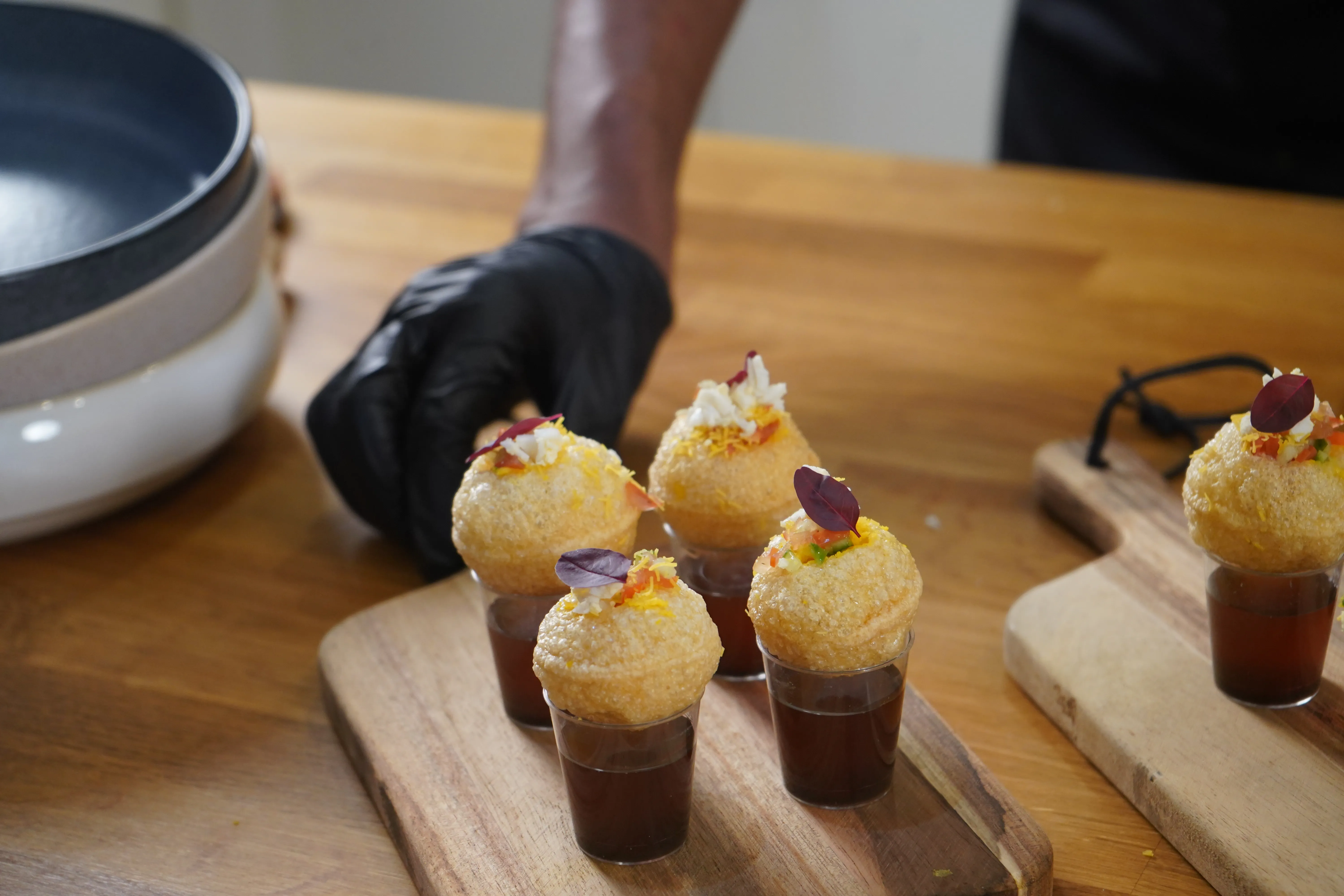 Gloved chef's hand placing a pani puri canapé on a wooden board.