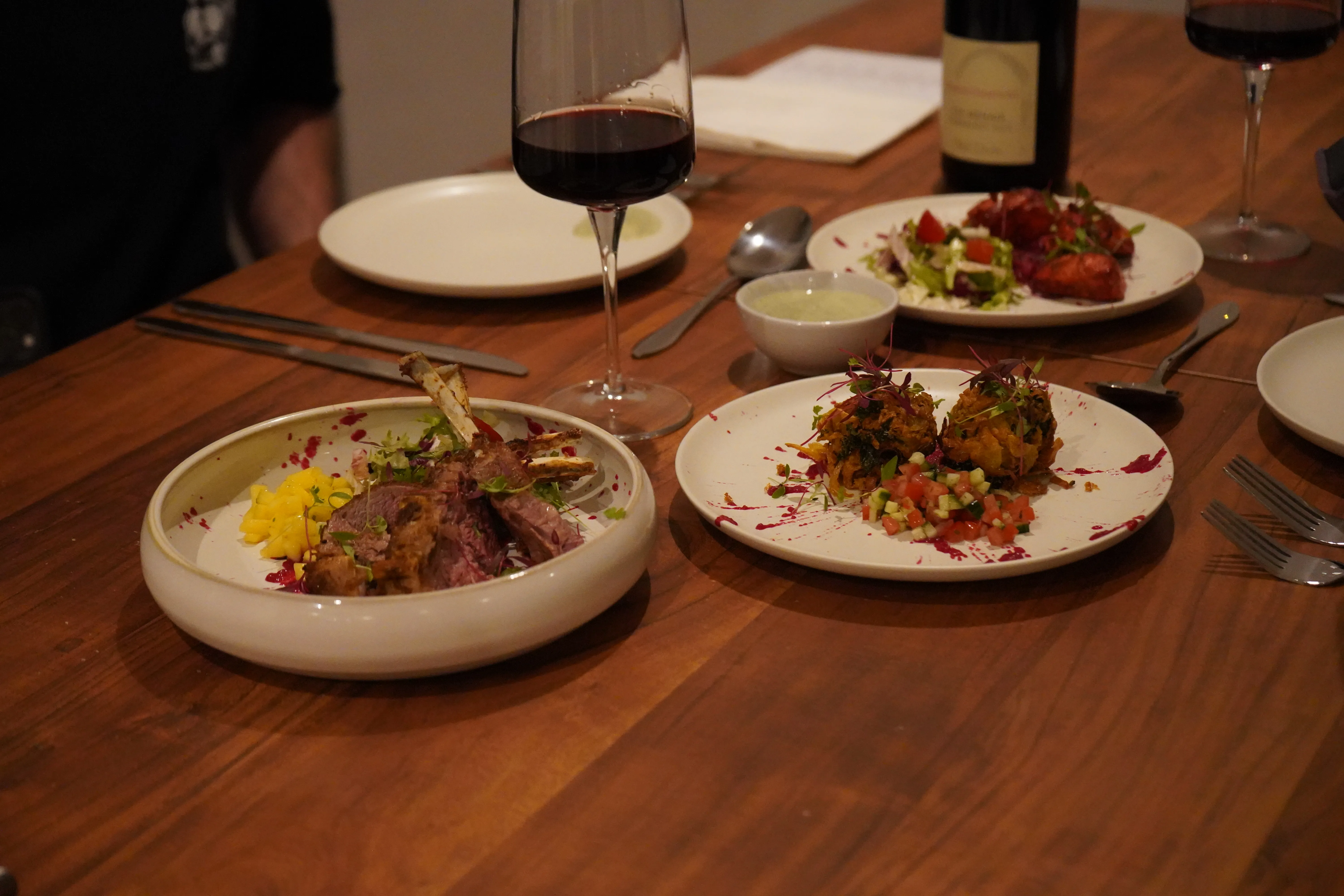 Warm wooden table set with lamb cutlets, mango chutney, bhaji and red wine.
